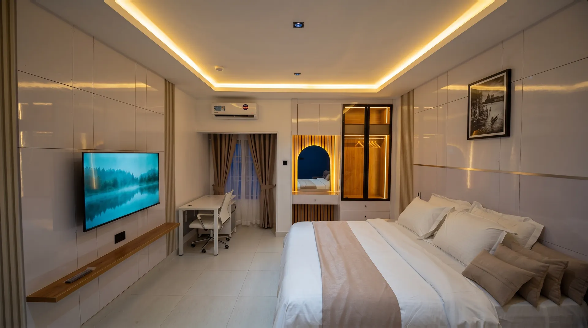 A bedroom in a Cholis Residency apartment in the evening: the bed made, a desk by the window, the television on and the wardrobe lit.